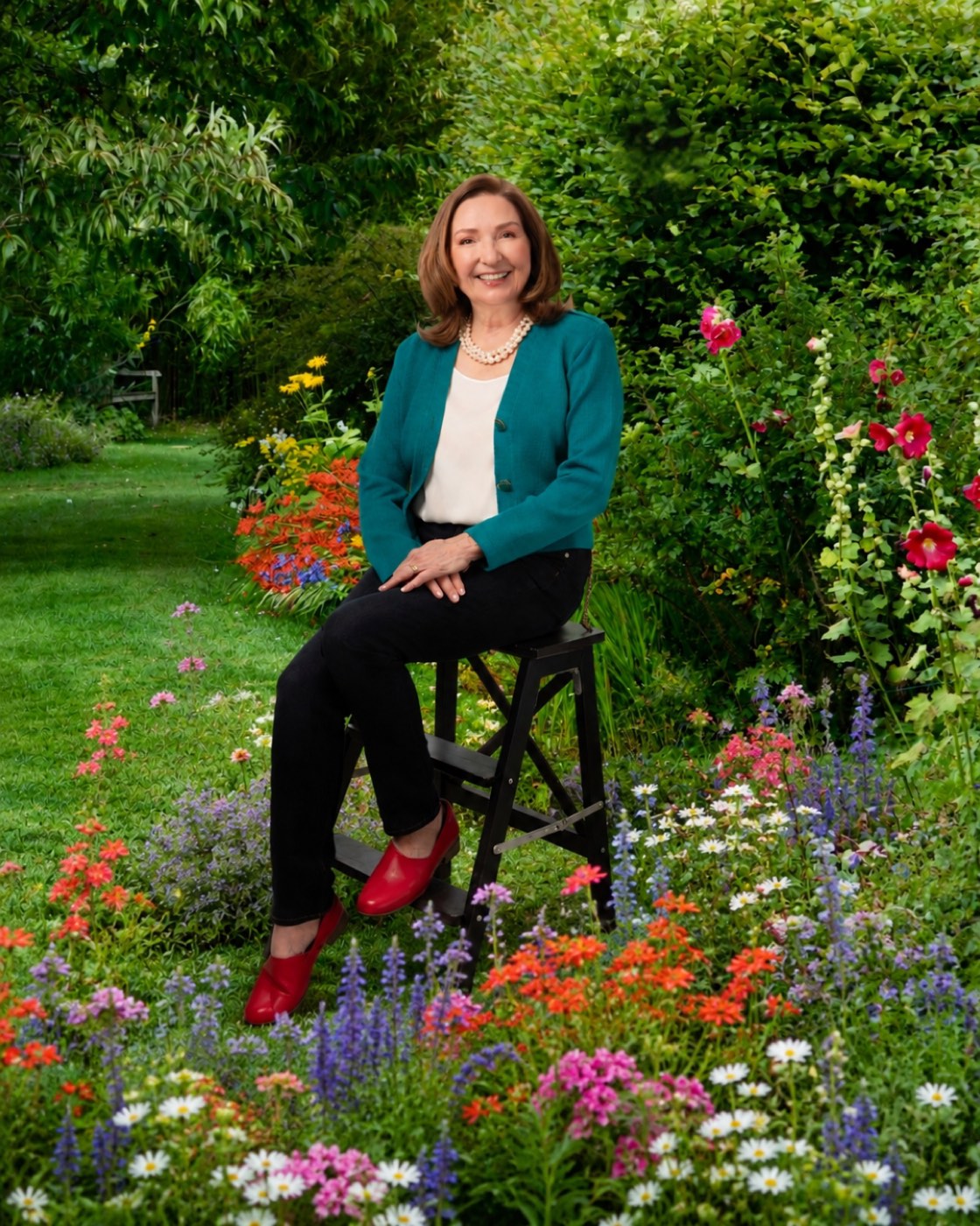 Portrait of author Ann Landers, LMFT, smiling in a teal cardigan, seated on a step stool in a flowering garden